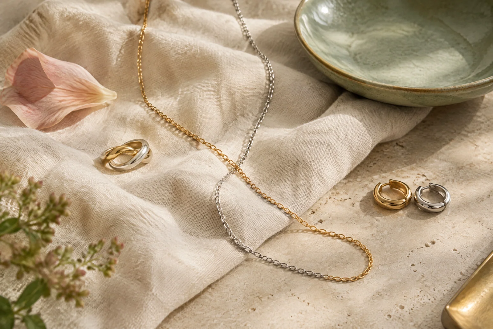 Gold and silver chains cross on cream linen beside a sage ceramic dish and soft blush petal.