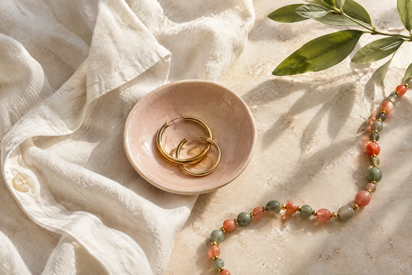 Conceptual gold-tone hoops and coral-sage beads arranged on white linen beside a blush ceramic dish.
