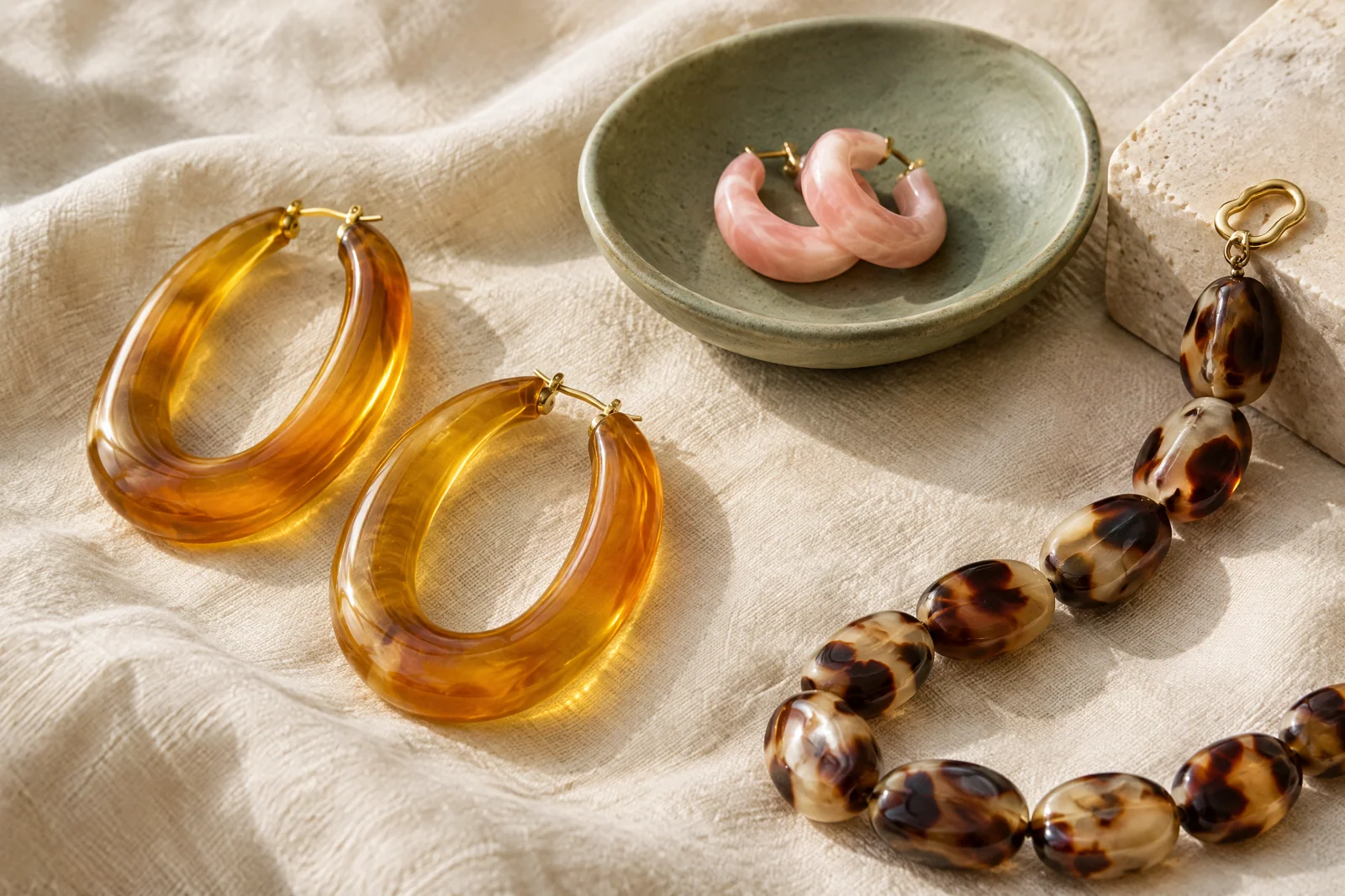 Translucent amber and blush resin hoops with patterned acetate beads arranged on cream linen beside sage ceramic and pale stone.